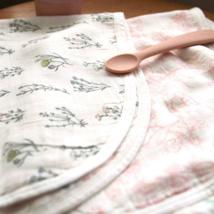 A pink baby spoon rests on a folded aden + anais boutique silky soft burpy bib, featuring white fabric with floral patterns. The bibs silky-soft texture and pastel colors create a gentle, cozy look.