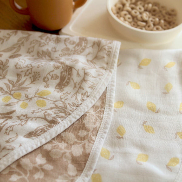 A close-up of two patterned cloths on a table showcases the boutique silky soft burpy bib 2 pack by aden + anais. One cloth features a floral and fruit motif in beige and white, while the other has a yellow lemon pattern. Nearby, a bowl of cereal and mug enhance this cozy scene.