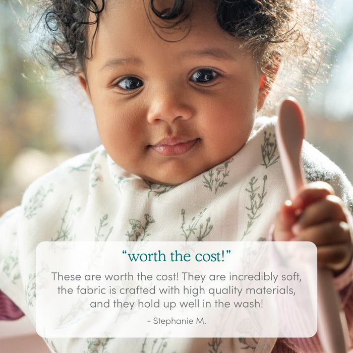 A curly-haired baby wears an aden + anais boutique silky soft burpy bib with a white and green leaf print, holding a pink spoon. Below, a review praises the 2 pack’s absorbency, high quality, and durability in the gentle sunlight.