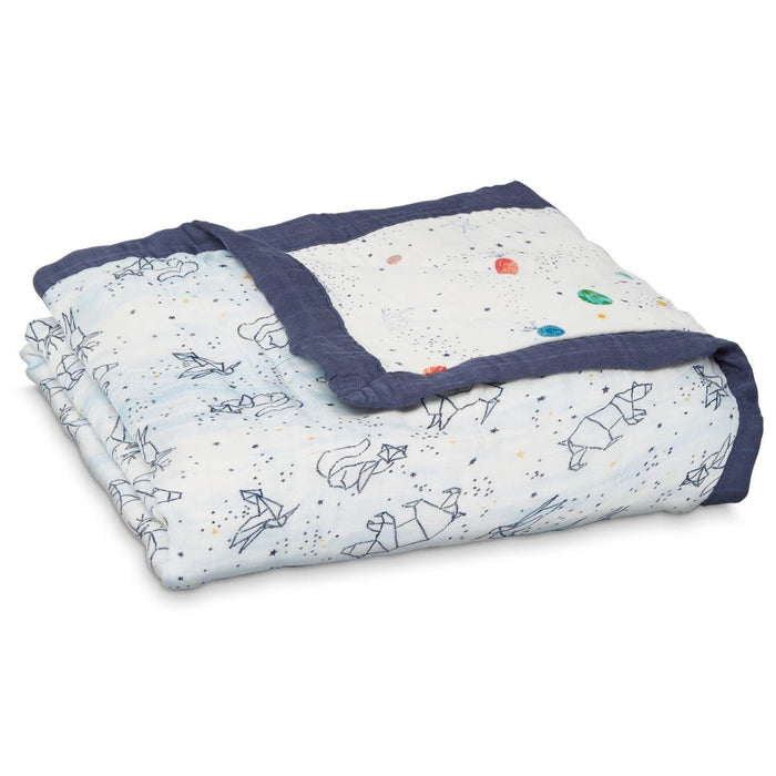 The aden + anais boutique silky soft muslin dream blanket is a cozy, folded white baby blanket with a dark blue border featuring space-themed rockets and planets in minimalistic lines and colorful dots. Crafted from bamboo-infused viscose, its perfect for wrapping your little one in comfort.
