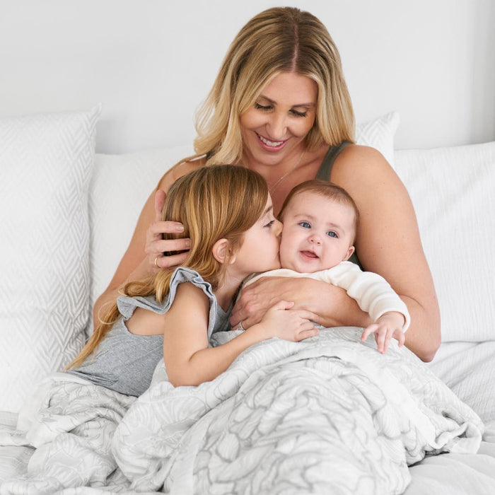 A woman smiles on a bed, holding a baby wrapped in an aden + anais boutique silky soft muslin dream blanket, while a young girl kisses the babys cheek in a bright, cozy setting with white pillows and bamboo-infused viscose bedding.