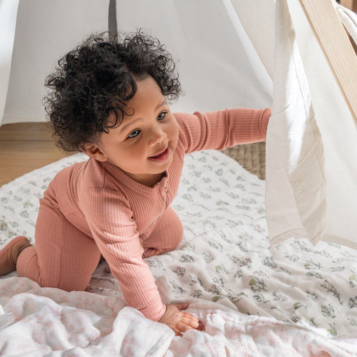A baby in pink pajamas lifts a curtain inside a tent, kneeling on an aden + anais boutique silky soft muslin dream blanket. The room is bright and cozy, with natural light illuminating the curly-haired babys cheerful expression as they beam in bamboo-infused viscose pajamas.