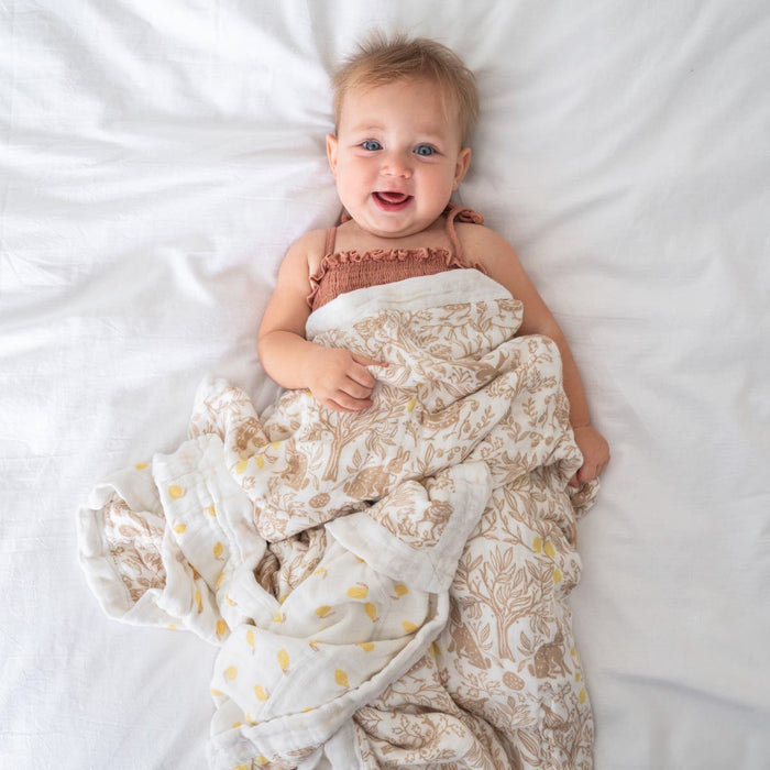 A smiling light-haired baby lies on a white bed, wrapped in the aden + anais Bamboo Baby Bundle with a cream and beige pattern, looking up happily and relaxed.