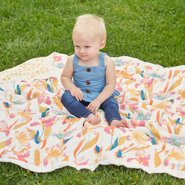 A baby in a blue top and jeans sits on an aden + anais boutique silky soft swaddle blanket with a colorful floral pattern, spread on green grass. The bamboo-infused viscose fabric adds smoothness as the baby gazes curiously to the side.