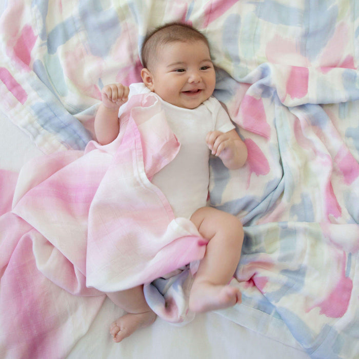 A baby in a white onesie lies on a bamboo-infused aden + anais boutique silky soft muslin dream blanket in shades of pink, blue, and white, smiling with arms slightly raised, surrounded by the cozy + cool fabric.