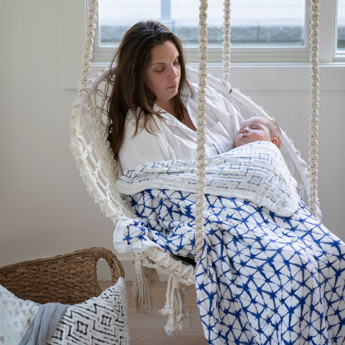 A woman sits in a cozy hanging chair, holding a sleeping baby wrapped in an aden + anais boutique silky soft muslin dream blanket, blue and white patterned. The room, with large windows behind her, features a bamboo-infused wicker basket on the floor. The ambiance is calm and soothing.