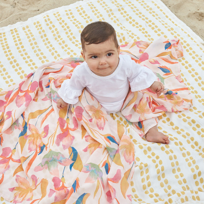 A baby with short dark hair sits on a sandy beach, wrapped in an aden + anais boutique silky soft swaddle blanket made from ultra-smooth, bamboo-infused viscose. The floral fabric covers a polka-dotted blanket as the baby wears a white outfit and looks upward.