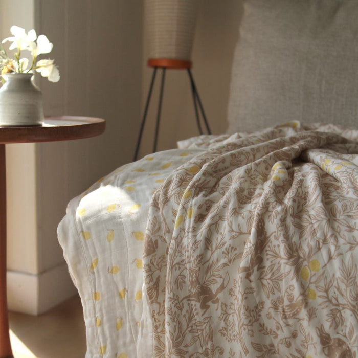 A cozy bed is adorned with the aden + anais boutique silky soft muslin dream blanket, showcasing light floral patterns and yellow dots. A wooden side table holds a vase with white flowers, while soft, natural lighting enhances the warm atmosphere.