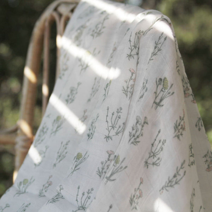 The aden + anais boutique silky soft muslin dream blanket, draped over a wooden chair, features a delicate floral pattern with green and soft pink accents. Sunlight streams through, creating cozy shadows on the blanket against a backdrop of blurred greenery.