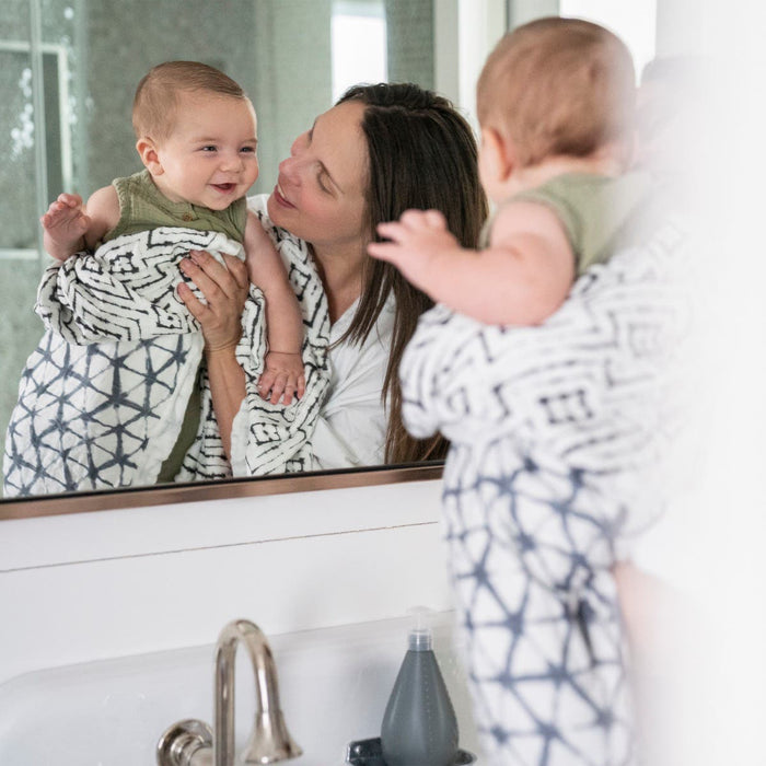 A woman joyfully holds a laughing baby swaddled in an aden + anais boutique silky soft muslin dream blanket, standing before a mirror in a bathroom with a visible sink and soap dispenser.