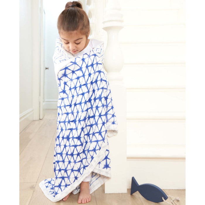 A young girl stands on a staircase wrapped in an aden + anais boutique silky soft muslin dream blanket. She looks down with a small blue toy fish beside her. The setting feels bright and airy, enhancing the cozy atmosphere.