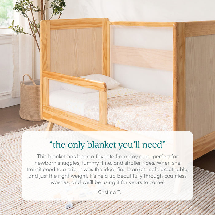 An aden + anais organic cotton muslin dream blanket, praised for its quality for newborns, is perfectly draped on a wooden crib with a light-colored mattress. A rug beneath enhances the rooms warmth, while a nearby plant adds greenery.
