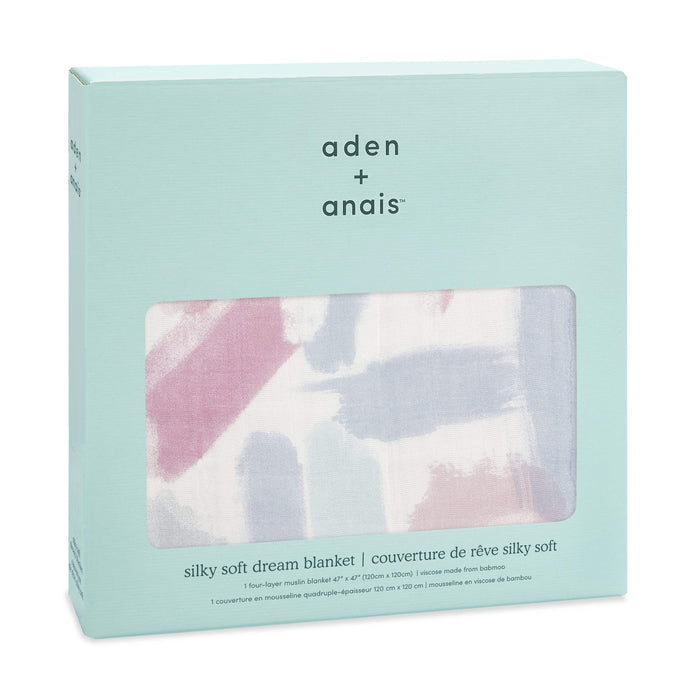 A light blue box displays a boutique silky soft muslin dream blanket by aden + anais, made from bamboo-infused viscose with abstract pink, blue, and gray swirls. The cozy and cool fabric is visible through the packaging window.