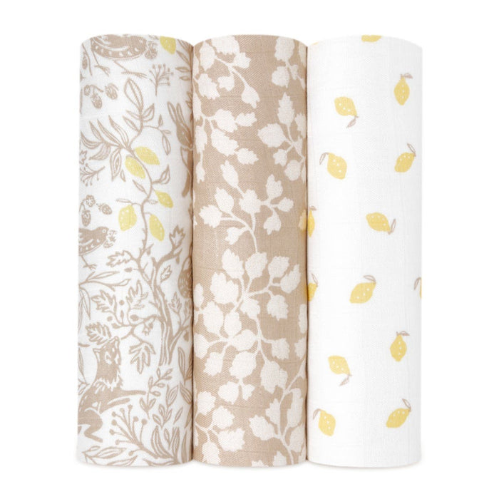 Three rolls of fabric stand upright: the left displays a bamboo-infused viscose with gray and yellow floral and animal patterns, the middle has a beige background with white leaves, and the right features an aden + anais boutique silky soft swaddle blanket on a white backdrop with yellow leaves.