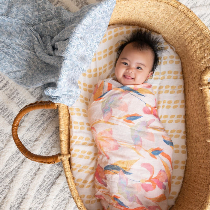 A baby is smiling while wrapped in a boutique silky soft swaddle from aden + anais, lying in a wicker basket. A patterned blue cloth lines the basket, which sits on a gray and white textured surface.