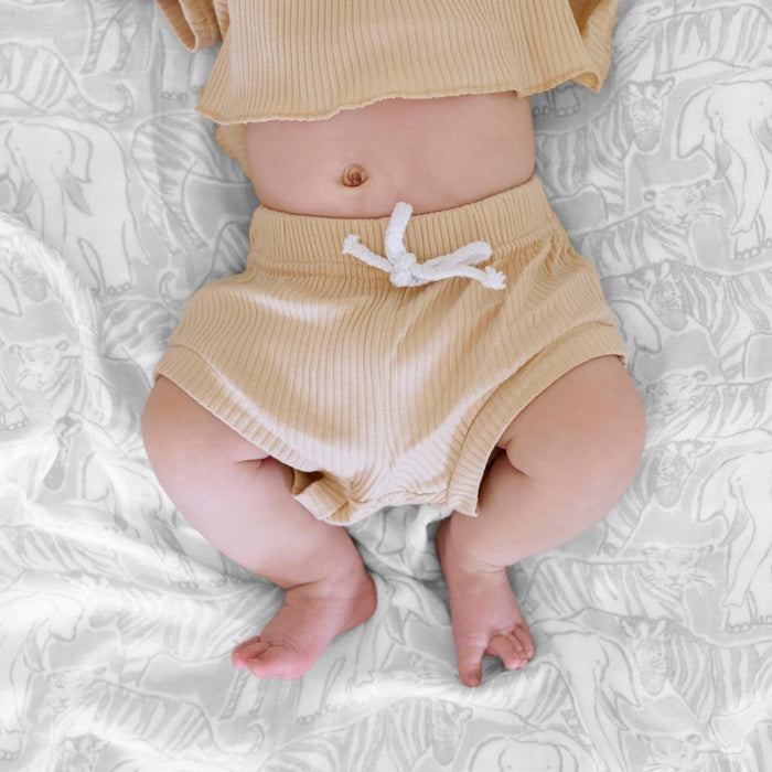 A baby in beige ribbed shorts with a white drawstring and matching top lies comfortably on gray and white elephant-patterned sheets, showcasing their belly button. The bamboo-infused viscose fabric of their outfit ensures silky soft comfort, similar to the aden + anais boutique swaddle blankets.