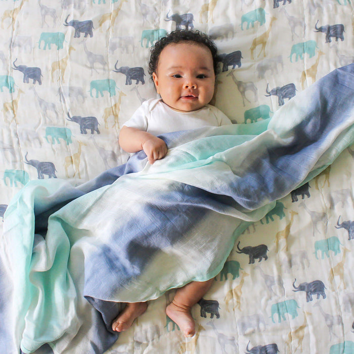 A baby on a bed covered with an elephant-patterned sheet is partly wrapped in an aden + anais boutique silky soft swaddle blanket, featuring a blue and white tie-dye design made of bamboo-infused viscose. The baby wears a white onesie, looking up playfully.
