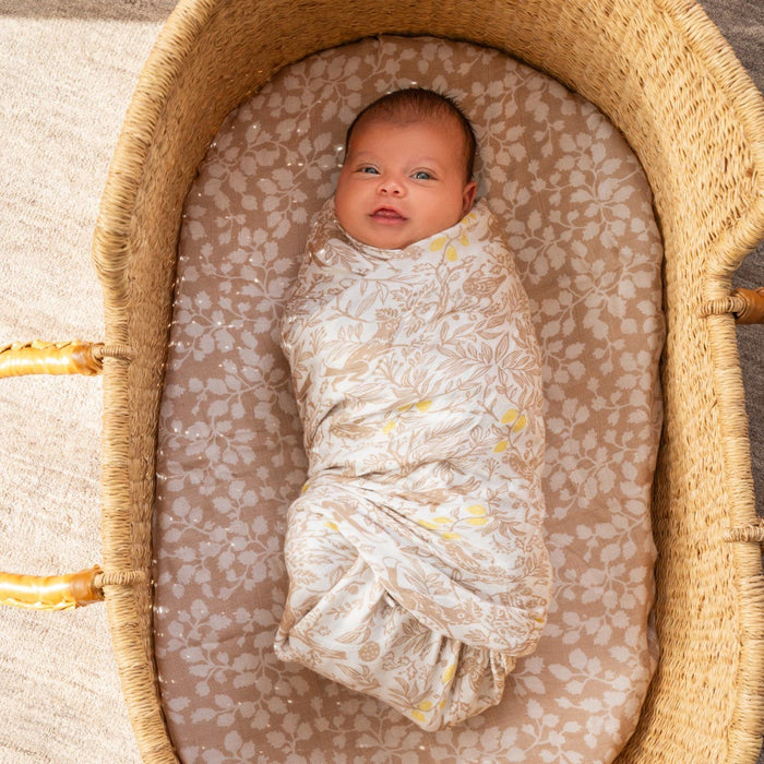 A baby wrapped in an aden + anais boutique silky soft swaddle blanket from the 3 pack, featuring floral patterns, lies comfortably in a woven bassinet with neutral-toned bedding.