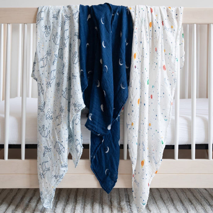Three aden + anais boutique silky soft swaddles are draped over a light-colored crib, featuring patterns of animal outlines, moons on a dark blue background, and colorful dots. A striped rug lies beneath.