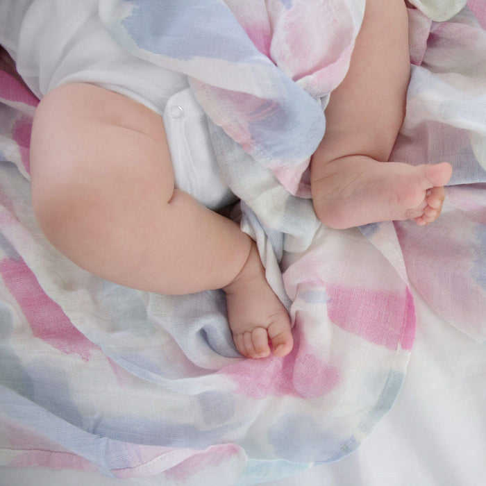 A baby in a white onesie is cozily wrapped in an aden + anais boutique silky soft swaddle blanket featuring pastel pink, blue, and gray patterns. The bamboo-infused viscose ensures breathability as the babys legs and feet rest gently on the white surface.