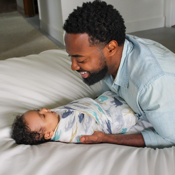 A man in a light denim shirt smiles at a baby swaddled in an aden + anais boutique silky soft swaddle blanket, lying on a white bed. The bamboo-infused viscose blanket makes them both look comfortable and content.