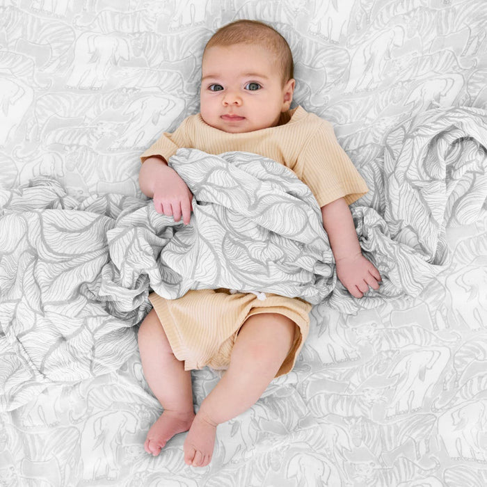 A baby lies on a patterned blanket, wrapped in an aden + anais boutique silky soft swaddle blanket from the 3 pack. The infant gazes up with wide eyes, tiny hands grasping the breathable swaddle amid a gentle gray and white leaf design that enhances the serene scene.