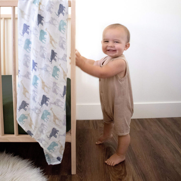 A toddler in a beige romper smiles beside a wooden crib with a dinosaur-patterned blanket draped over it. The cozy room, featuring wooden flooring and a white wall, is accentuated by aden + anais boutique silky soft swaddle blankets 3 pack made of bamboo-infused viscose neatly tucked in.