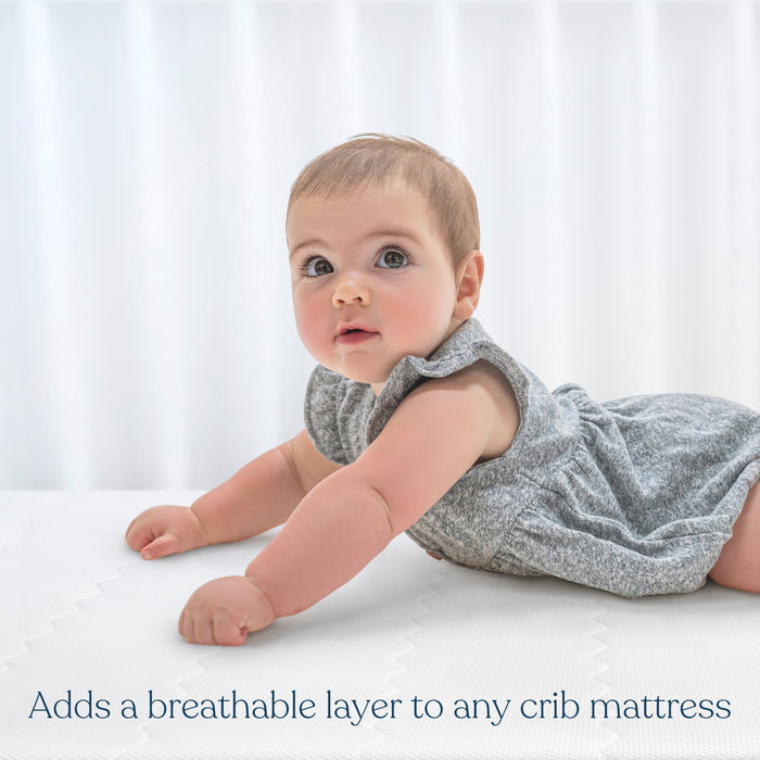 A baby in a gray outfit lies on a crib mattress with HALOs Breathable Crib Mattress Cover. Resting on their stomach, the baby gazes upward as white curtains frame the scene. Text reads, Adds a breathable layer to any crib mattress.