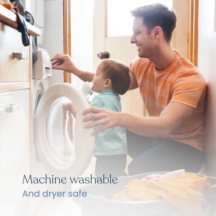 In a laundry room, a man helps a child open the front-loading washing machine. On the floor lies a basket of clothes beside HALOs Breathable Crib Mattress Cover with waterproof lining. Its labeled Machine washable and dryer safe.