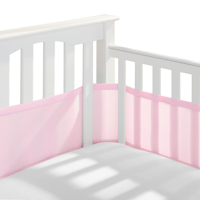 A white crib with slatted sides displays attached light pink HALO Breathable Mesh Crib Liners for a full-sized crib. Built to meet infant bedding safety standards, the empty crib shows gentle sunlight shadows on the white mattress.