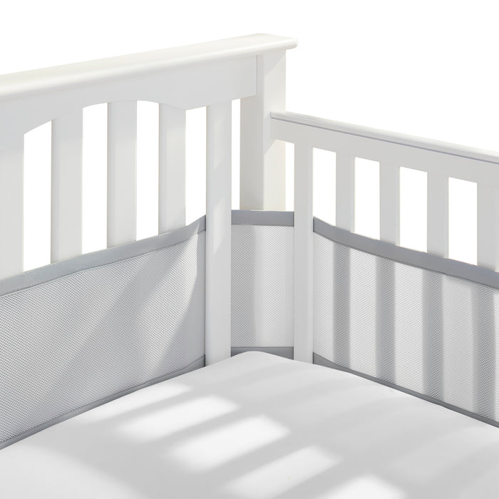 Close-up of a white baby crib corner with slatted sides, featuring the HALO Breathable Mesh Crib Liner for Full Sized Crib in light gray. The mattress, under a white sheet, lies serenely beneath soft shadows.