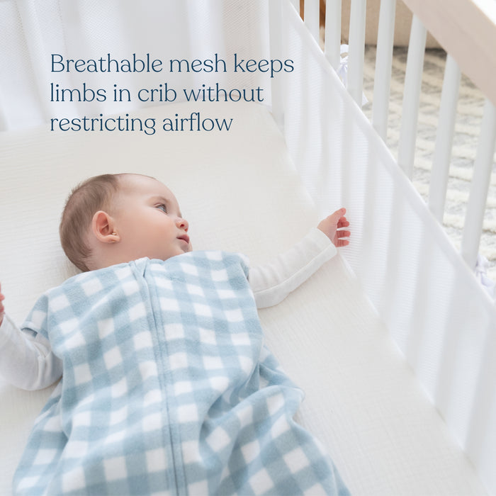 A baby in a blue and white checkered sleep sack lies in a crib with the HALO Breathable Mesh Crib Liner for Full-Sized Crib, ensuring safety by keeping limbs inside and allowing airflow. Text on image: Breathable mesh keeps limbs in crib without restricting airflow.