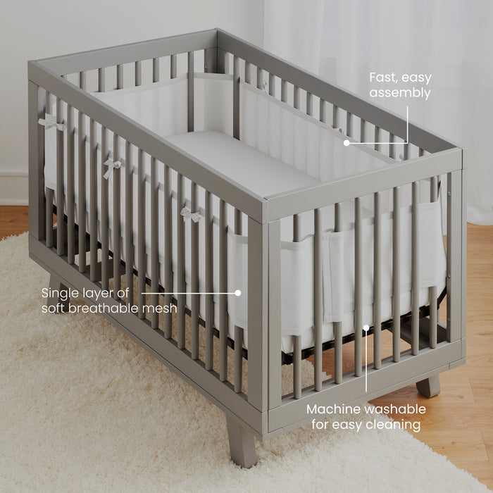 A gray crib with the HALO Breathable Mesh Crib Liner is set against a white curtain, featuring quick assembly and safety features like soft, machine washable parts. An infant-themed cream rug lies beneath.
