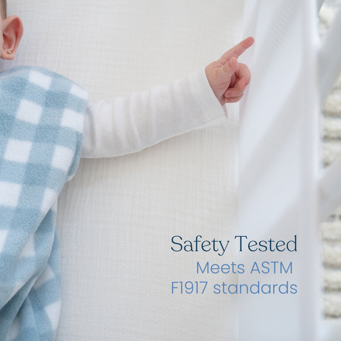 A baby sleeps peacefully in a crib wrapped in a blue and white checkered blanket, with its tiny hand pointing upwards. The text stresses Safety Tested Meets ASTM F1917 standards, underscoring infant safety with the HALO Breathable Mesh Crib Liner for Full Sized Solid End Cribs.