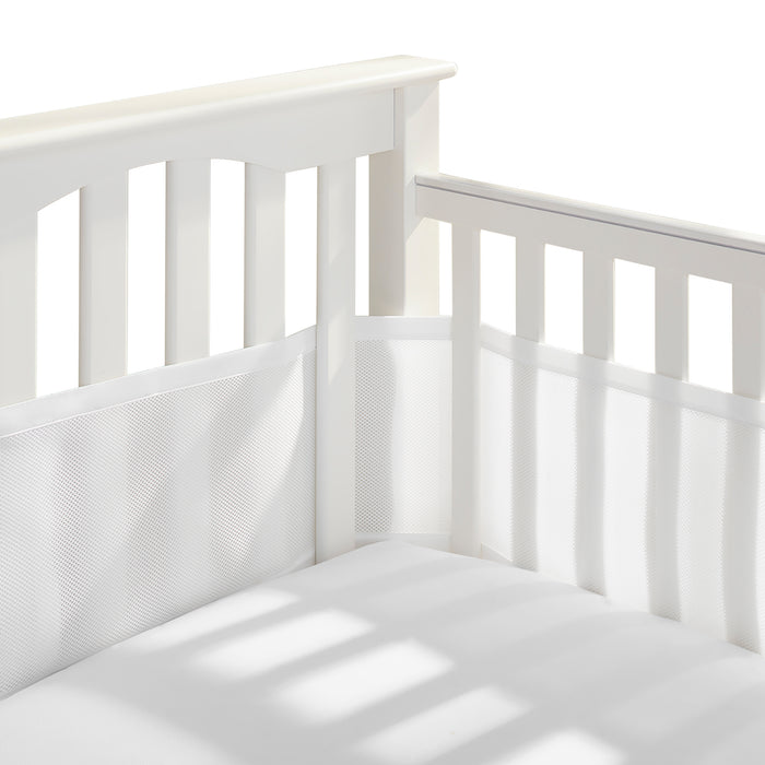 Close-up of a white crib corner showing vertical slats with a HALO Breathable Mesh Crib Liner for Mini Crib. Its minimalist design ensures safety, casting soft shadows on the infant bedding and liners.