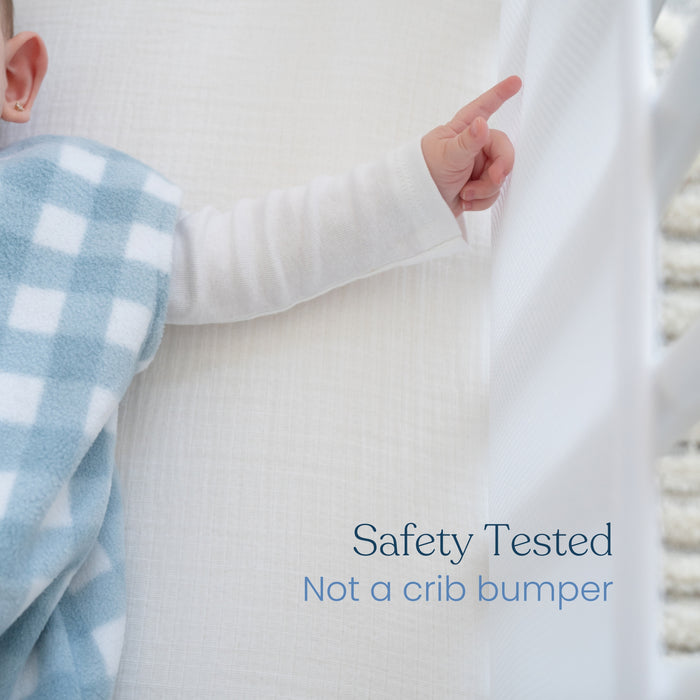 A babys arm in a blue checkered outfit is raised next to a white crib rail, showcasing HALOs Safety Tested Breathable Mesh Crib Liner for Full Sized Crib, featuring enhanced crib safety with Not a crib bumper clearly labeled for peace of mind.