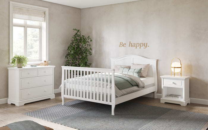 A bright, cozy bedroom features the Child Craft Camden 4-in-1 Convertible Crib in matte white, a matching dresser and nightstand, a green plant by the window, and "Be happy." on the wall, with soft colors and sunlight creating a peaceful mood.
