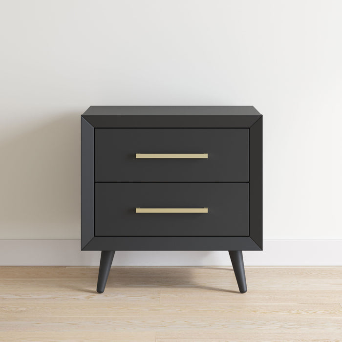 The Child Craft Cranbrook Nightstand, Ebony, is a modern black nightstand with two drawers, metallic handles, and four angled legs, shown against a white wall on a light wood floor.