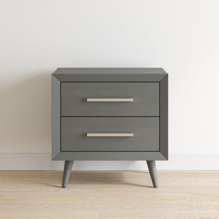The Child Craft Cranbrook Nightstand in Lunar Gray features two drawers with silver handles and stands on angled legs, set against a white wall on a light wood floor.