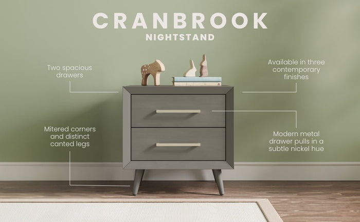 The Cranbrook Nightstand by Child Craft in Lunar Gray features two drawers with metal handles and stands on angled legs. Displayed against a green wall, it blends modern style with functionality, as shown with decorative items on top.