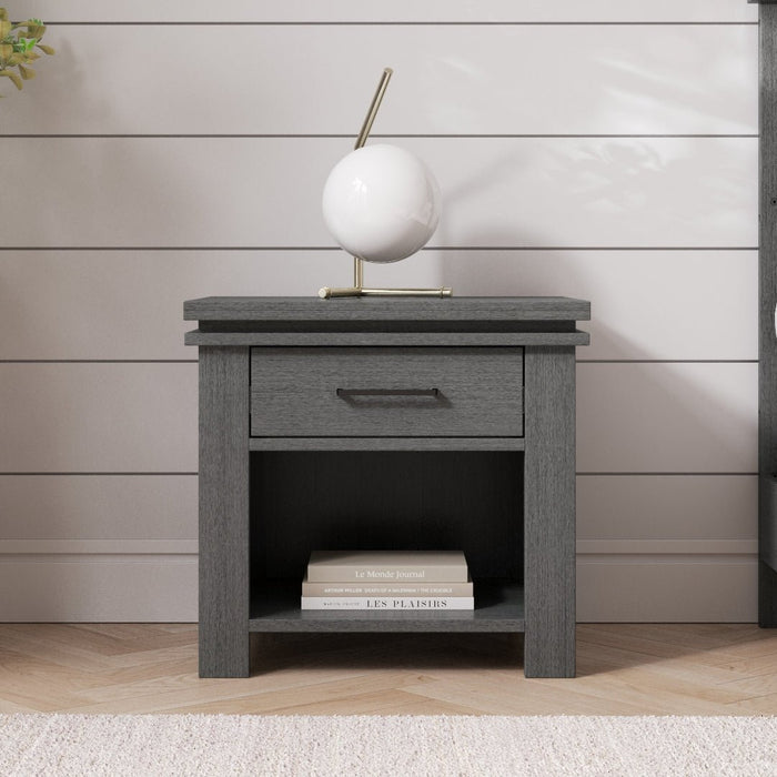 The Child Craft Denman Nightstand in Midnight Gray features a drawer and open shelf for books, topped with a modern white and gold lamp, set against a white shiplap wall on a light wooden floor with a rug.