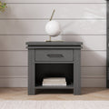 The Child Craft Denman Nightstand in Midnight Gray features a drawer and open shelf for books, topped with a modern white and gold lamp, set against a white shiplap wall on a light wooden floor with a rug.