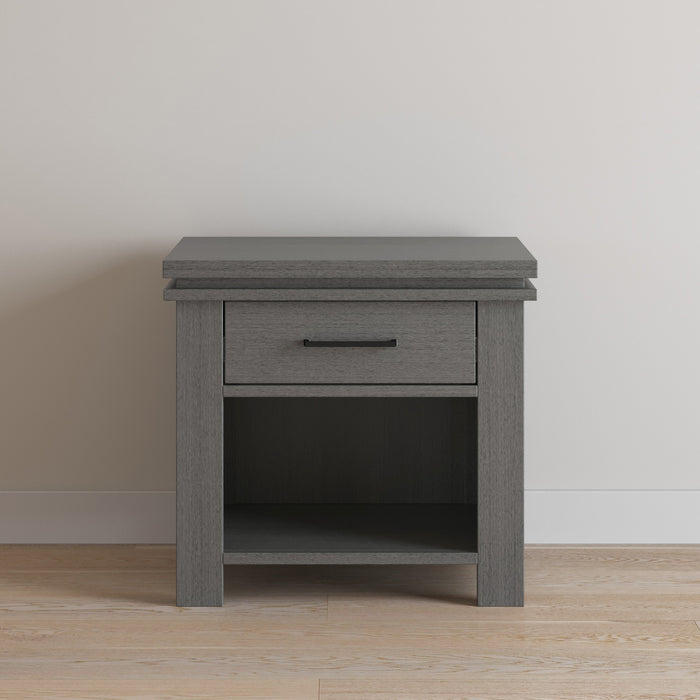 The Child Craft Denman Nightstand in Midnight Gray features a single drawer and open lower shelf, shown against a light wall on a light wood floor.