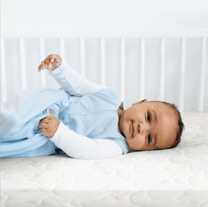 A baby in a light blue outfit lies on a mattress fitted with the HALO DreamWeave Breathable Crib Cover in a white crib. Resting on their side, the infant looks at the camera. The cribs white rails and soft lighting create a peaceful atmosphere, enhanced by its GREENGUARD Gold certification.