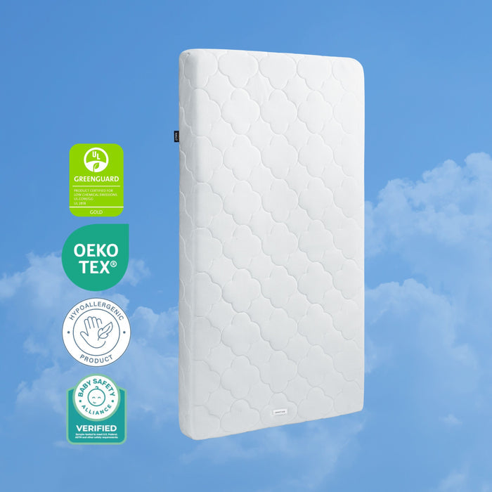 The HALO DreamWeave™ Breathable Crib Mattress stands upright against a blue sky with clouds, displaying four badges: GREENGUARD Gold, Oeko-Tex, Hypoallergenic Product, and Baby Safety Alliance Verified.