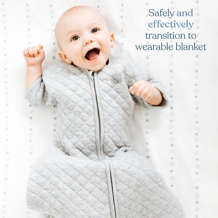 A baby smiles while lying on a white surface, wrapped in HALOs Easy Transition SleepSack TOG 1.5, a gray quilted wearable blanket. Text reads: Safely transition to our 3-layer design for better sleep.