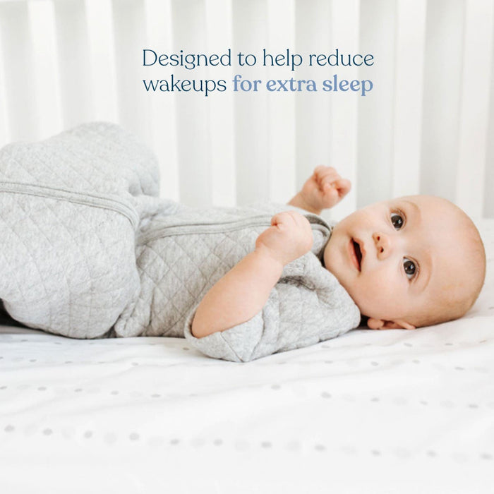 A baby rests on the Easy Transition SleepSack TOG 1.5 by HALO, cozy in gray quilted fabric. The infant smiles slightly, framed by white crib bars. Text above reads, 3-layer design for better sleep and easy transition—helping reduce wakeups for extra sleep.