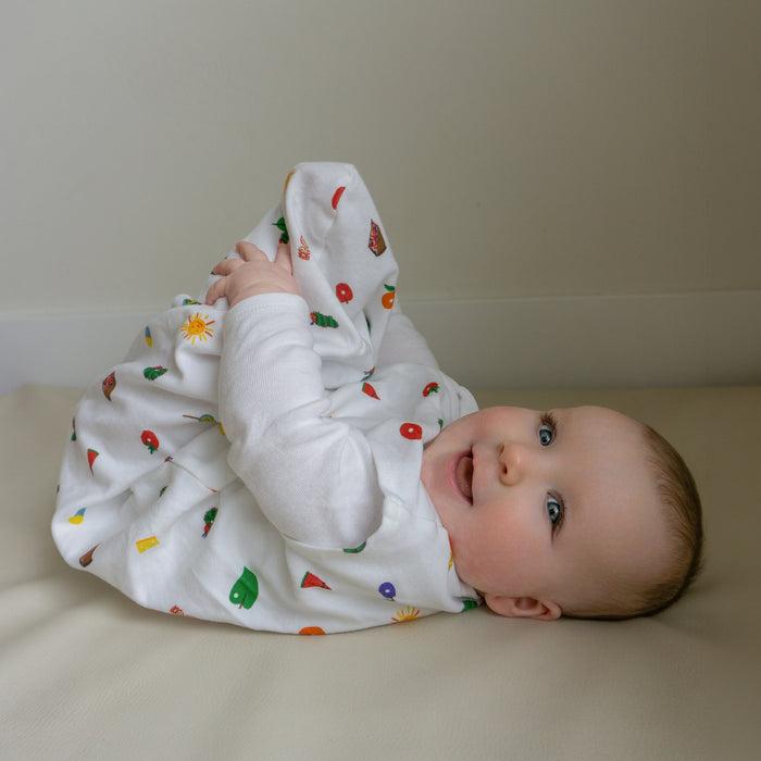 A baby smiles and holds their feet while lying on their back in a HALO Eric Carle The Very Hungry Caterpillar 100% Cotton Wearable Blanket TOG 0.5, featuring colorful fruit and vegetable prints.