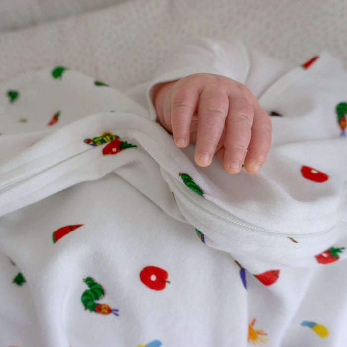 A baby’s hand rests on a white HALO Eric Carle The Very Hungry Caterpillar 100% Cotton Wearable Blanket TOG 0.5 decorated with colorful fruit and caterpillar designs.