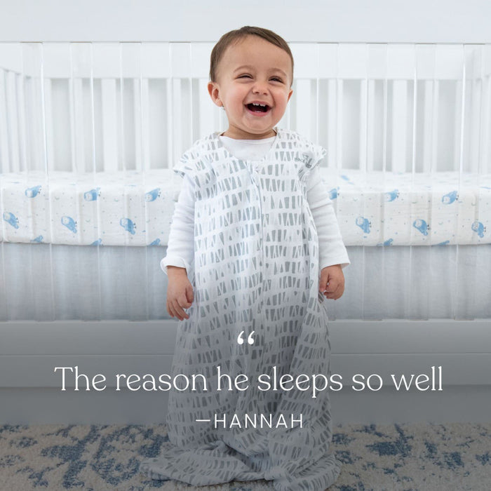A smiling baby stands in a HALO Eric Carle The Very Hungry Caterpillar 100% Cotton Wearable Blanket (TOG 0.5) in front of a white crib with blue and white bedding. Text reads: “The reason he sleeps so well — Hannah.”.